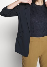 Námořnický modrý blejzr s klopami, lehká tkanina a přední kapsa, spárovaný s námořnickým krajkovým topem a světle hnědými kalhotami s vysokým pasem.