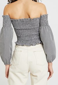 Bluse med off-the-shoulder design, smocket liv, ballonærmer i et sort-hvidt gingham mønster, flæse i kanten, kombineret med lyse jeans.