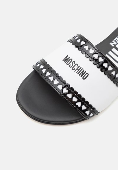 Sandale à talon plat noire et blanche avec une sangle brillante comportant des découpes en forme de cœur et le logo « MOSCHINO », semelle plate et texture lisse.