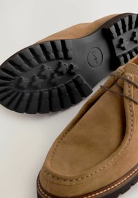 Mocassins en daim marron avec détails cousus, semelle extérieure en caoutchouc noir flexible présentant un motif de bande de roulement, et accents en lacet sur le dessus.
