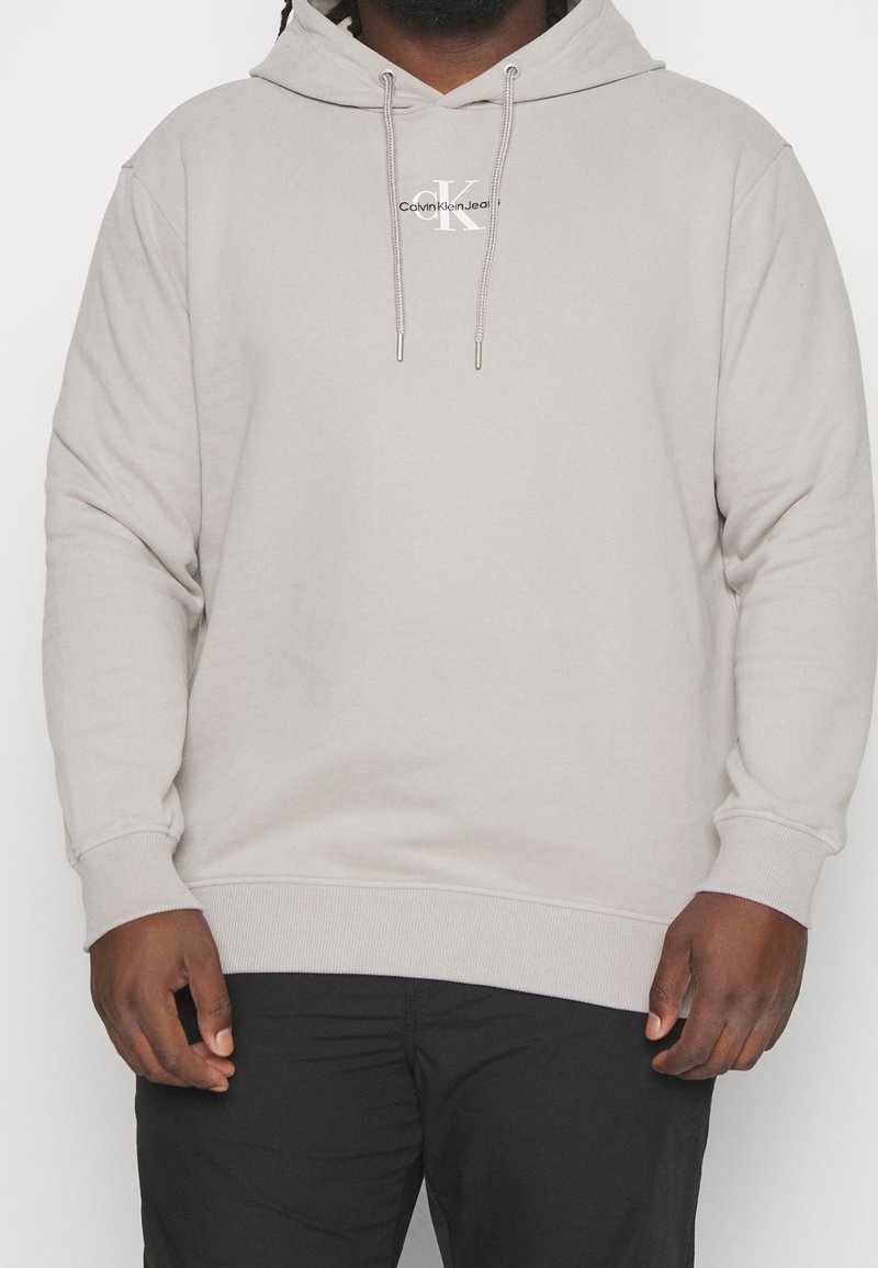 Homme portant un sweat à capuche Calvin Klein beige clair avec cordons et pantalon noir, visible des épaules aux hanches.
