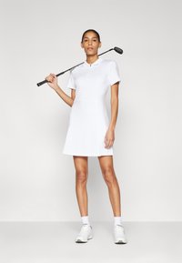 Bela golf obleka s kratkimi rokavi, ovratnikom, oprijetim pasom in razširjenim krilom. Model drži golf palico in nosi bele superge.