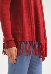 Main portant un pull rouge à manches longues avec un ourlet à franges, reposant à côté d'un pantalon bleu clair.