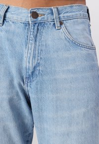 Blugi din denim de un albastru deschis, cu talie înaltă și fermoar, având buzunare frontale și detalii discrete de estompare pe întreaga țesătură.