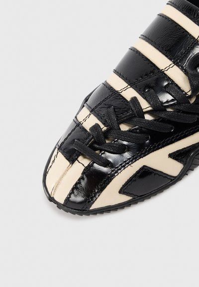 Black and beige sneaker with a glossy finish, featuring horizontal stripes, textured surface, and a lace-up design. Notable side accents present.