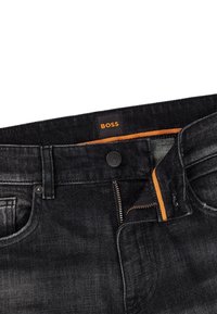 Jeans de mezclilla oscura con una cinturilla negra, costuras naranjas y un cierre frontal. Presenta una etiqueta de marca con "BOSS" en color naranja.