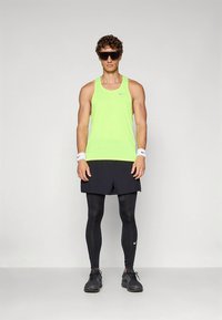 Neonowo zielony sportowy tank top, czarne szorty, czarne legginsy i czarne buty sportowe. Dodatki obejmują białe opaski na nadgarstki i okulary przeciwsłoneczne.