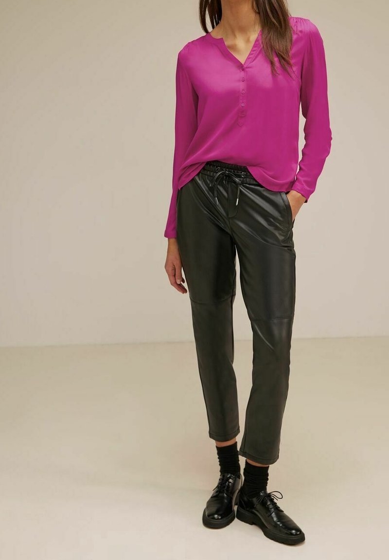 Femme portant un chemisier rose vif à manches longues, un pantalon noir en simili cuir avec taille à cordon, des chaussettes noires et des chaussures noires à lacets.