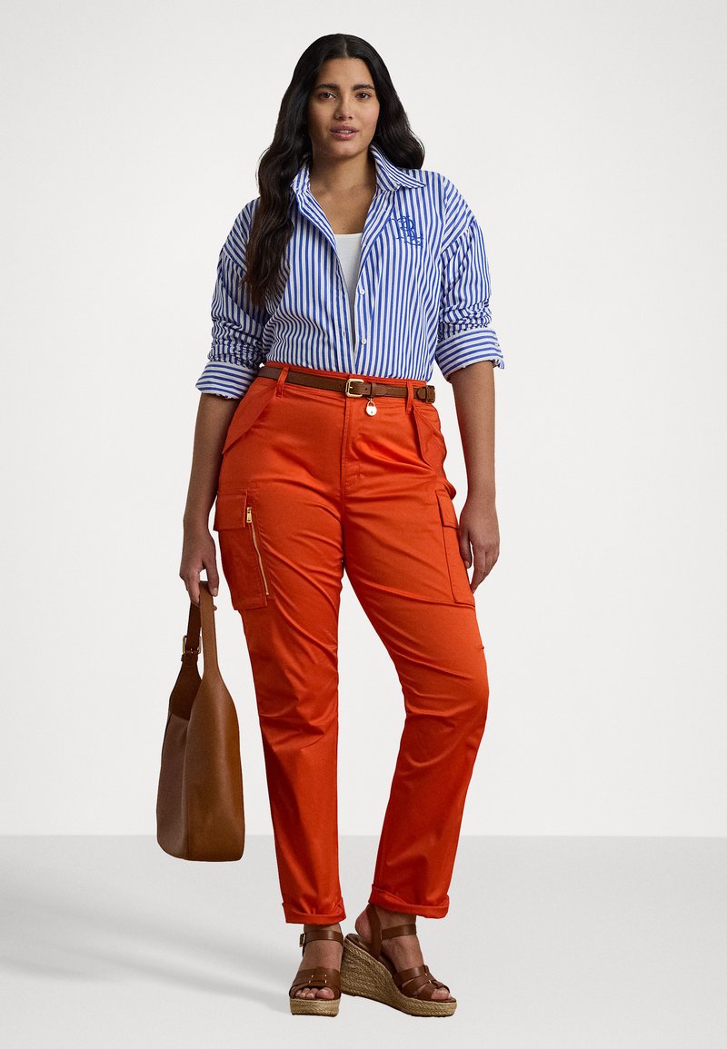 Camisa a rayas azul y blanca, pantalones cargo naranjas con bolsillos laterales y un cinturón marrón. Bolsa de hombro marrón y sandalias de cuña.