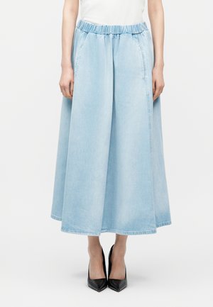 Γυναίκα που φοράει ανοιχτό μπλε τζιν κουλοτσές με φαρδιά μπατζάκια και ελαστική μέση, σε συνδυασμό με μαύρες ψηλοτάκουνες γόβες με μυτερή μύτη, στέκεται μπροστά σε λευκό φόντο.