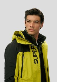 Water-resistant jacket in bright yellow and black. Features a zipper closure, adjustable hood, and large logo print on the front.