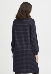 Marinekleid mit grünem Karomuster, langen Ärmeln mit elastischen Bündchen und geradem Schnitt. Verfügt über eine Rückennaht und keine sichtbaren Verschlüsse.