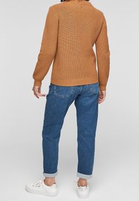 Gebrande oranje gebreide trui met geribbelde textuur, hoge kraag en logo-detail op de rug, gecombineerd met blauwe opgerolde jeans en witte sneakers.