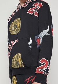 Sudadera con capucha negra con estampados gráficos coloridos, que incluyen insignias metálicas doradas y motivos de toros. Cuenta con un bolsillo frontal y forro de capucha a rayas.