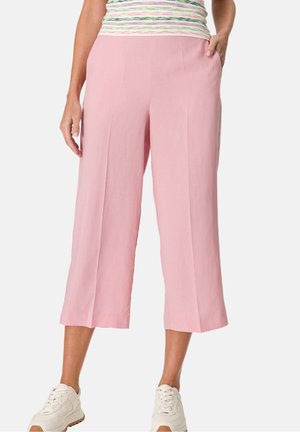 Une femme portant un pantalon cropped large rose clair et des baskets blanches, avec une main dans sa poche, devant un fond uni.