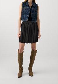 Džínová vesta bez rukávů, spárovaná s černou skládací sukni a hnědými kozačkami ke kolenům se špičatými špičkami a bočními zipy.