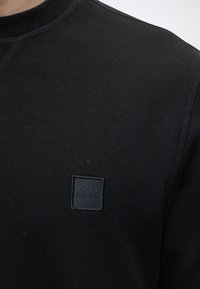 T-shirt noir à col rond avec un petit patch carré portant le logo "BOSS" sur la partie supérieure gauche de la poitrine.