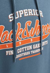 Modrá bavlnená mikina s kapucňou s výrazným bielym a oranžovým potlačeným textom, vrátane "Superior" a "Jack & Jones," s kapucňou na šnúrku.