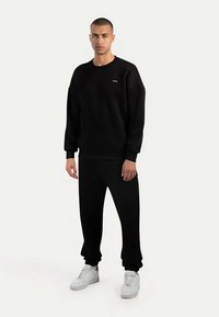 Completo con felpa nera e pantaloni jogger in tessuto morbido, caratterizzato da scollo rotondo e polsini a coste, abbinato a scarpe da ginnastica bianche.