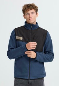 Fleece bunda v modrej farbe s hornou časťou z čierneho nylonu, vysokým golierom, zipsom a vreckom na hrudi. Na hrudi je umiestnené logo.