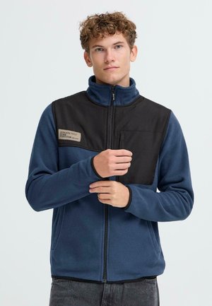 Chaqueta de forro polar en azul con sección superior de nylon negro, cuello alto, cierre de cremallera y bolsillo en el pecho. Logo destacado en el área del pecho.