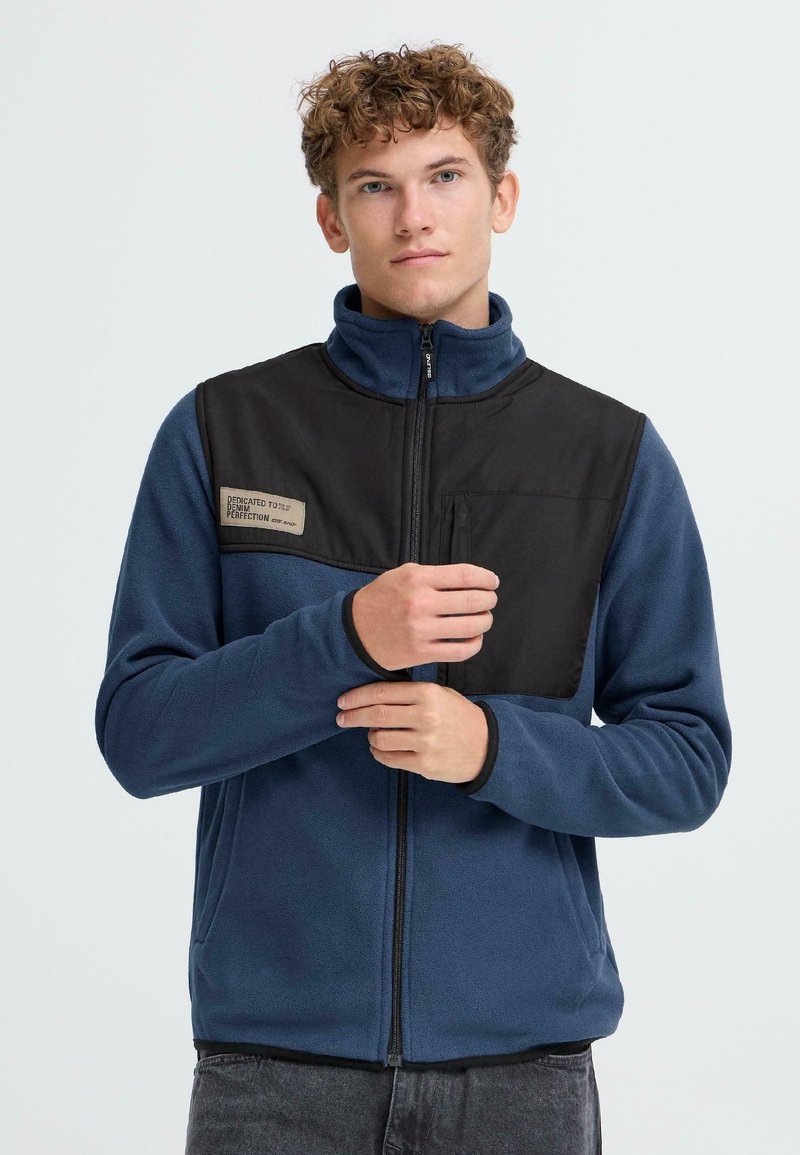 Fleece bunda v modrej farbe s hornou časťou z čierneho nylonu, vysokým golierom, zipsom a vreckom na hrudi. Na hrudi je umiestnené logo.