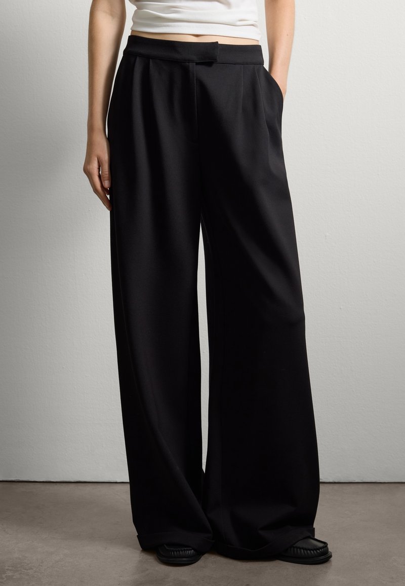 Pantalones anchos negros con una textura suave, cintura, bolsillos laterales y un corte relajado, combinados con calzado negro.