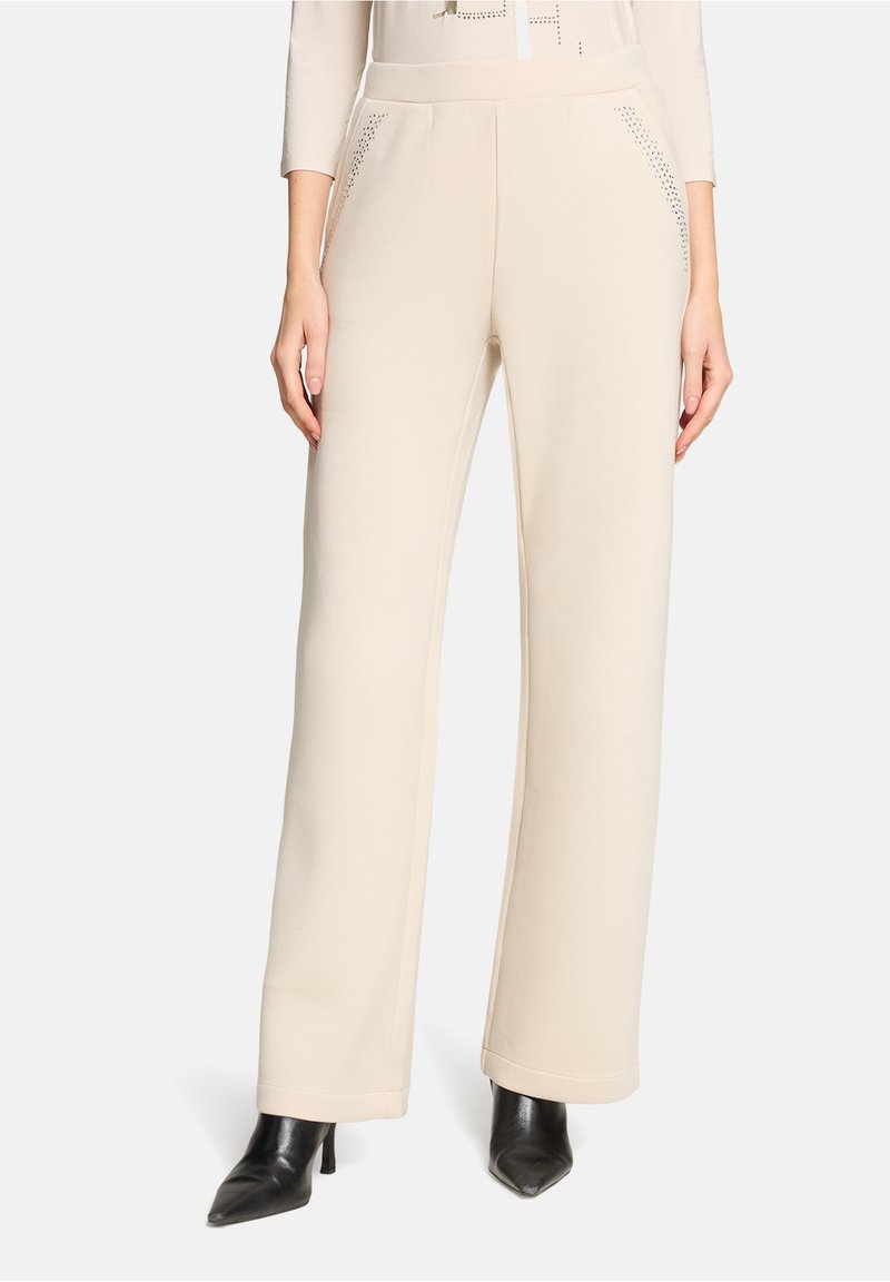 Pantalon beige à jambes larges avec une texture lisse, doté de poches latérales ornées de subtils clous, associé à des talons noirs à bout pointu.