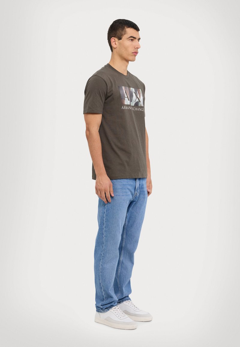 T-shirt vert olive avec un logo "AX" imprimé, associé à un jean droit bleu clair et des baskets blanches avec des accents gris.