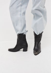 Bottes de cheville en cuir noir avec un bout pointu et un accent argenté à la pointe, présentant un talon carré et une couture détaillée. Portées avec un pantalon bleu clair.