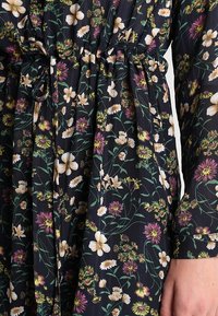 Flödig svart klänning med ett blommönster som har vita och lila blommor; dragsko i midjan, långa ärmar och lätt strukturerad stoff.