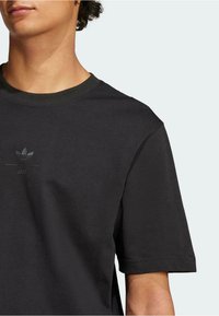 Črna bombažna majica s kratkimi rokavi in krožnim izrezom, ki ima vtisnjen logotip Adidas in besedo "SS25" pod njim. Gladka tekstura.