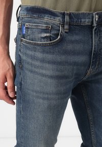 Jeans de denim en azul oscuro con un ligero desgaste, con un diseño clásico de cinco bolsillos, remaches metálicos y una etiqueta con el logo en el bolsillo trasero.