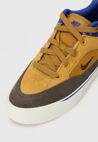 Adidași bej și maron cu un superior din ante și material textil, având accente albastre, o suprafață texturată și o talpă din cauciuc cu detalii albe.