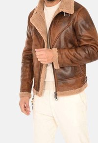 Mężczyzna noszący brązową kurtkę skórzaną z kożuchem w beżowym kolorze, w połączeniu z białą koszulką z okrągłym dekoltem i kremowymi spodniami.