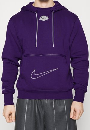 Homme portant un sweat à capuche violet des Lakers avec un logo Nike blanc et des cordons gris, assorti à un short noir avec des coutures blanches verticales.