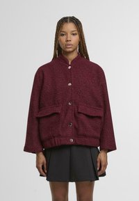 Donna che indossa una giacca testurizzata bordeaux con bottoni argentati abbinata a una gonna nera plissettata, in piedi su uno sfondo semplice.