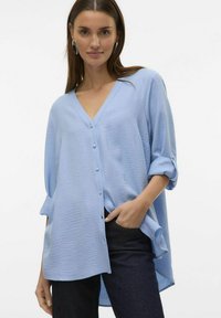 Lichtblauwe blouse met een V-hals, knoopsluiting en opgerolde mouwen. Heeft een textuurstof en een asymmetrische zoom. Gecombineerd met donkere jeans.