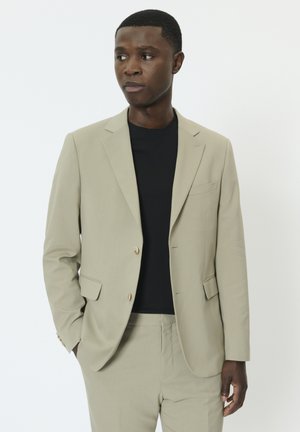 Világos bézs színű zakó, egysoros gombolású, két gombbal és hajtókás gallérral, fekete környakú pólóval párosítva. Sima anyagból.