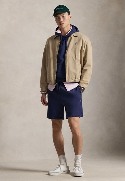 Smėlio spalvos striukė, tamsiai mėlynas džemperis su raišteliais, matosi rožinės spalvos marškinio apykaklė, tamsiai mėlyni šortai, balti sportbačiai ir žalia kepuraitė su logotipu.