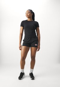 Camiseta negra de manga corta combinada con pantalones cortos negros que presentan un logo de Champion. El modelo lleva zapatillas negras y calcetines blancos de caña baja.