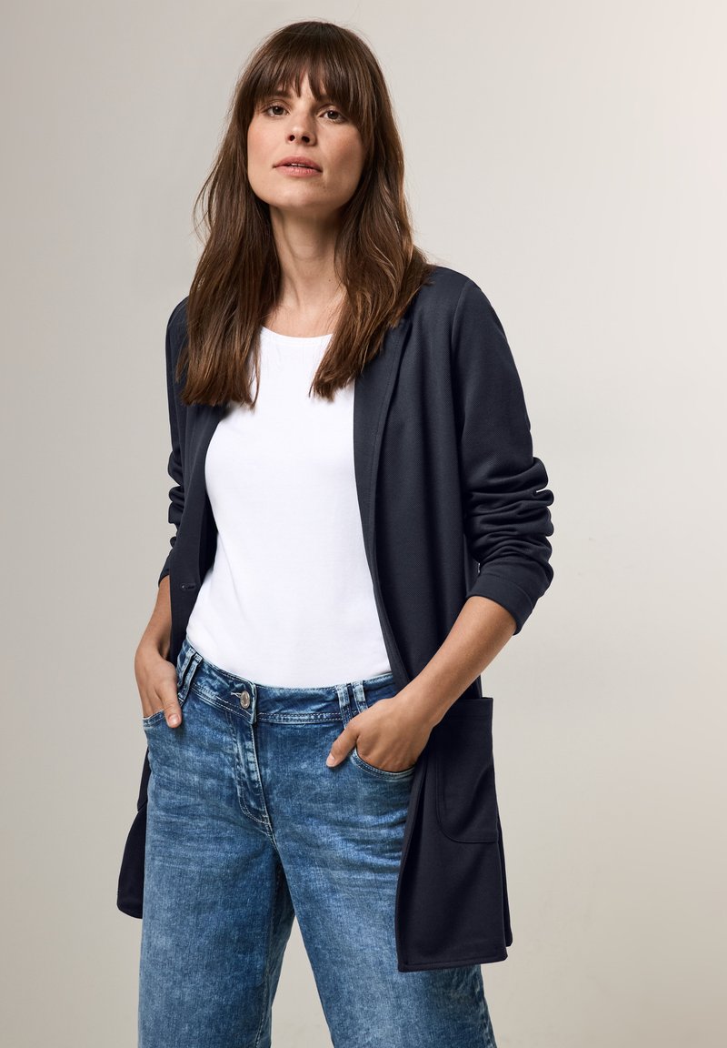 Cardigan lungo blu navy con maniche lunghe e tasche, indossato sopra una camicia bianca. Jeans di denim azzurro chiaro con una texture sbiadita e vestibilità standard.