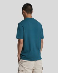 Tirkīzzila kokvilnas T-krekls ar apaļu apkakli un īsām piedurknēm; gluda tekstūra un brīvs piegriezums, valkāts ar gaiši bēšām kargo šortiem.