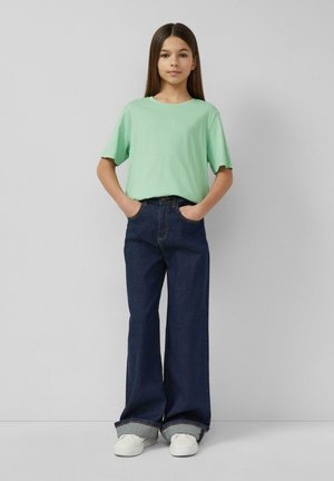 Fille debout, les mains dans les poches, portant un t-shirt vert menthe, un jean large bleu foncé avec le bas retroussé et des baskets blanches.