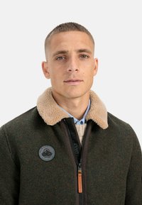 Green wool jacket with a tan fleece collar, black zip, and round patch. Features a textured finish and a zippered closure.