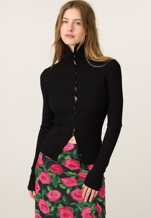 Vrouw die een zwarte geribde trui met knopen en een roze-groene bloemenrok draagt, staand tegen een effen lichte achtergrond.
