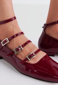 Burgunderfarbene Lacklederschuhe mit mehreren dünnen Riemen und silbernen quadratischen Schnallen. Spitz zulaufende Schuhform und niedriger Absatz. Glatte, glänzende Textur.