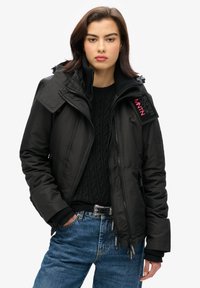 Czarna, ocieplana kurtka z odpinanym kapturem, z wysokim kołnierzem, zapięciem na zamek oraz widocznymi naszywkami marki. W połączeniu z czarnym wełnianym swetrem.