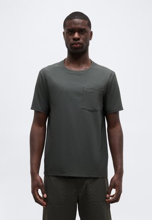 Jeune homme debout, faisant face à l'avant, portant un t-shirt à manches courtes gris foncé avec une poche poitrine et un pantalon foncé assorti.