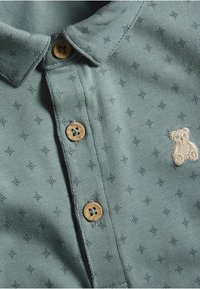 Polo-paita haaleassa vihreässä puuvillassa, jossa on pieniä tummia kuvioita, kaksi beigeä nappia ja brodeerattu nallekarhu-yksityiskohta rinnassa.